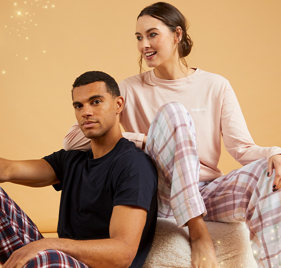 A man and woman sit on a fluffy stool against a peach background. Both are wearing plaid pajamas.