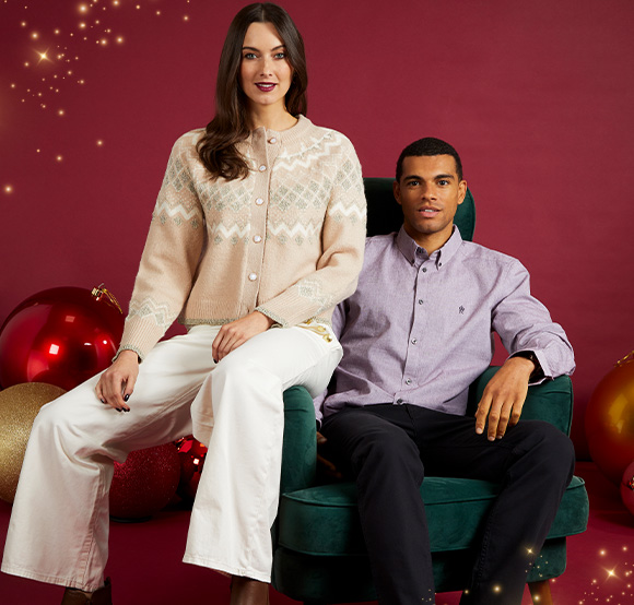 A woman in a beige sweater and white trousers sits on the arm of a green chair, next to a man in a purple shirt. They pose against a red backdrop with oversized Christmas ornaments.
