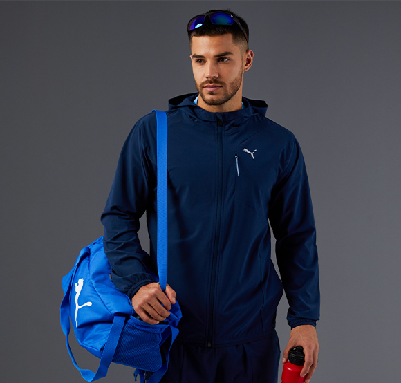 Male wearing a navy blue Puma hooded jacket, carrying a blue Puma sports bag and a red water bottle.