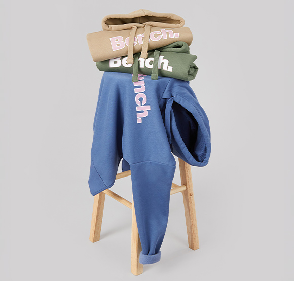 Three Bench hoodies in beige, olive green and blue, stacked and draped over a wooden stool.